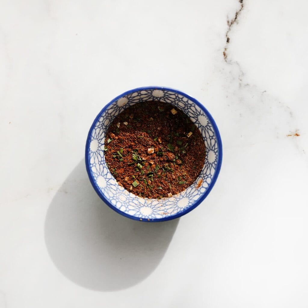 Meng voor het kruidenmengsel de knoflookpoeder, uienpoeder, cayennepeper, zwarte peper, korianderpoeder, paprikapoeder, komijnpoeder, gedroogde peterselie, kaneelpoeder en tijm in een kom.