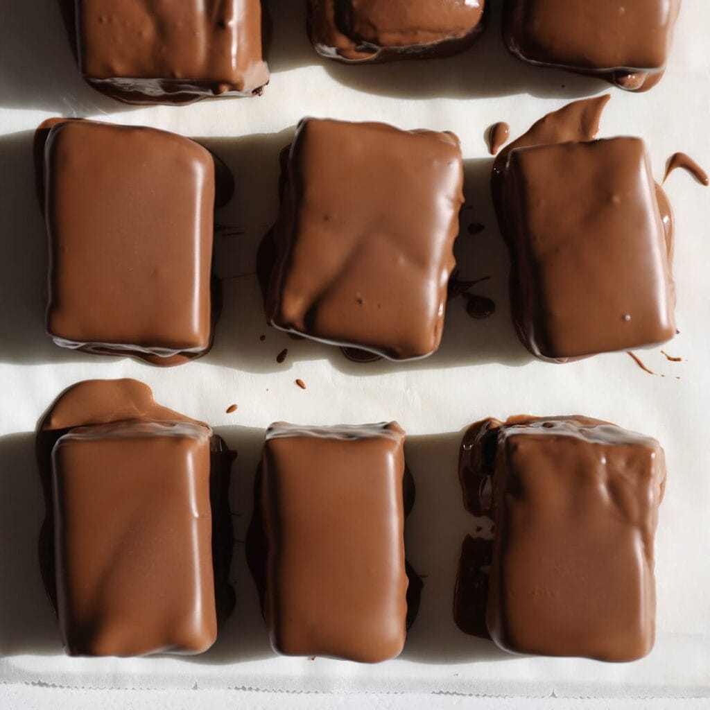 Snijd de cake in stukken. Haal elk stuk door de gesmolten melkchocolade en leg op bakpapier. Spuit er dunne lijnen pure chocolade overheen. Laat de chocolade uitharden.